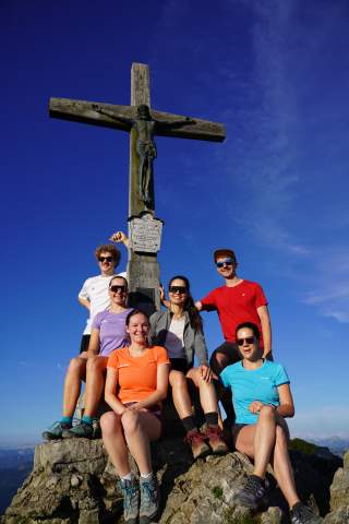 Sechs Personen in bunter Wanderkleidung stehen und sitzen unter einem Gipfelkreuz aus Holz mit einer Jesus-Figur und lachen in die Kamera, im Hintergrund blauer Himmel