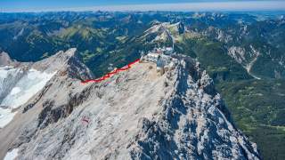 Luftaufnahme eines felsigen Berggipfels mit Gebäuden und einer roten Markierung, die einen Wanderweg zeigt, im Hintergrund grüne Täler und weitere Bergketten.